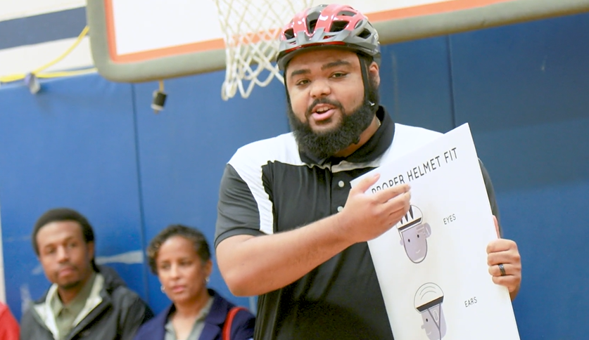 PE teacher speaking and holding sign that says "proper helmet fit."