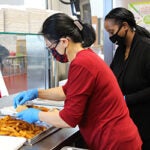 Two cafeteria supervisors prepare meals for students in a school lunch room.