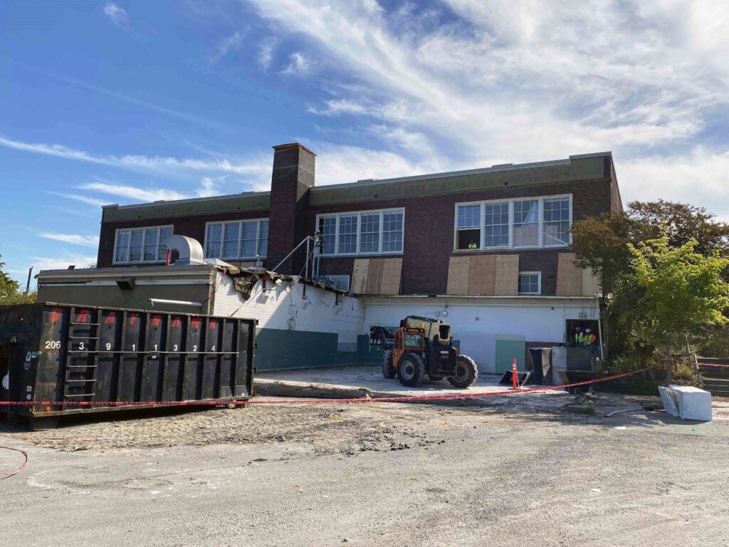 a two story brick building has windows on the top floor. an excavator is sitting below it on a concrete pad with only two walls remaining. a dumpster is next to the concrete