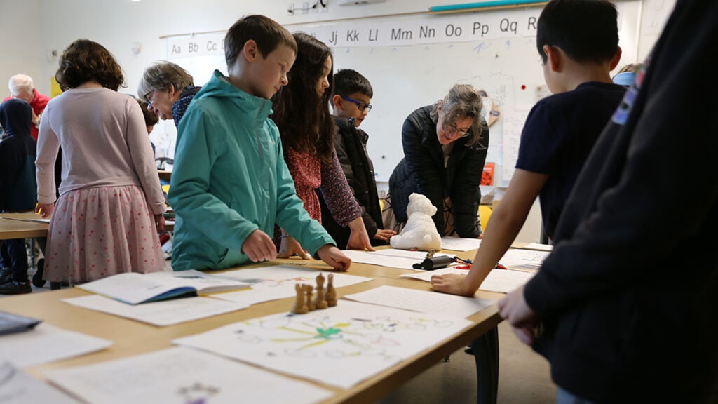 Students and elders stand around desks covered in artifacts, discuss objects.