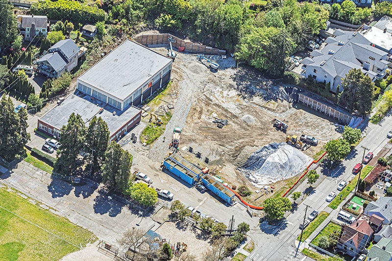 Aerial view of a construction site with a building in the upper left and construction equipment next to a partially built retaining wall