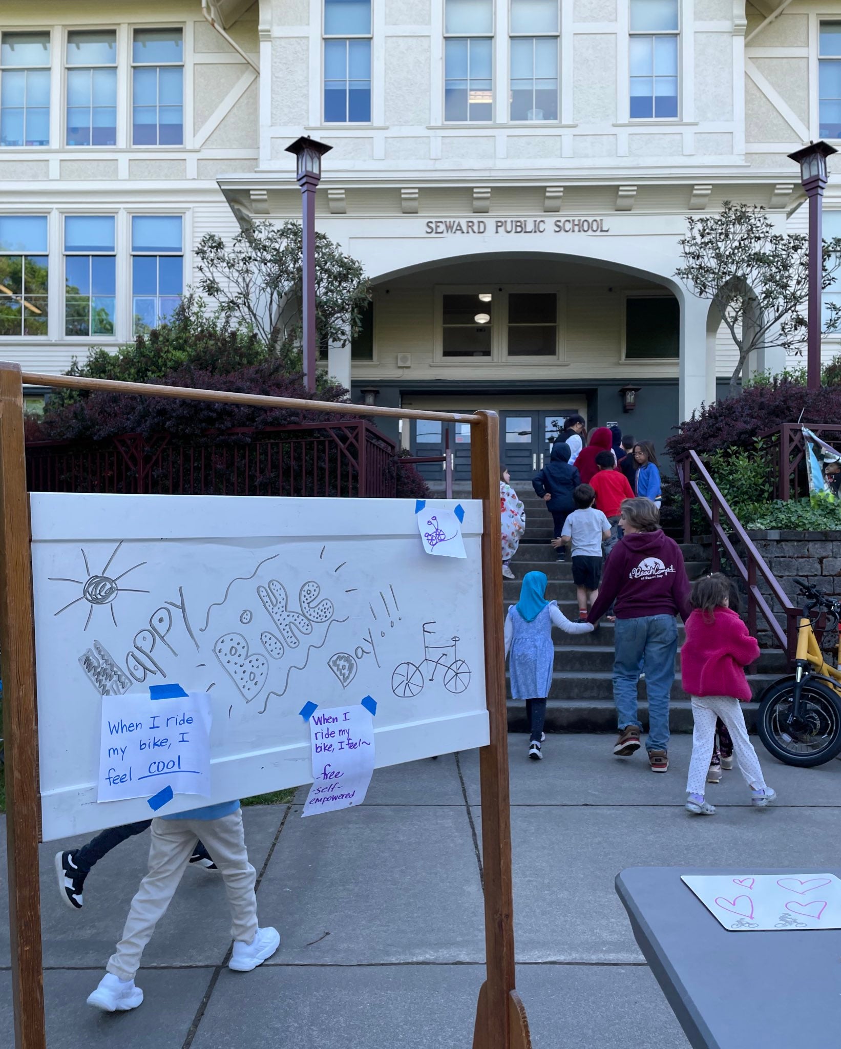 Sign in front of school says "Happy Bike Month" and features student notes. Student walk up steps into school in the background.
