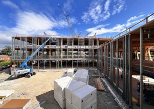 exterior of a large building under construction with walls partially framed, a person on a lift near the 3rd floor, construction materials in foreground and bright blue sky with light clouds.