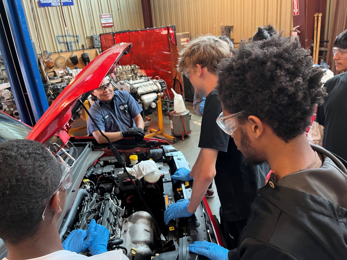 Three students looking at car with hood popped and teacher