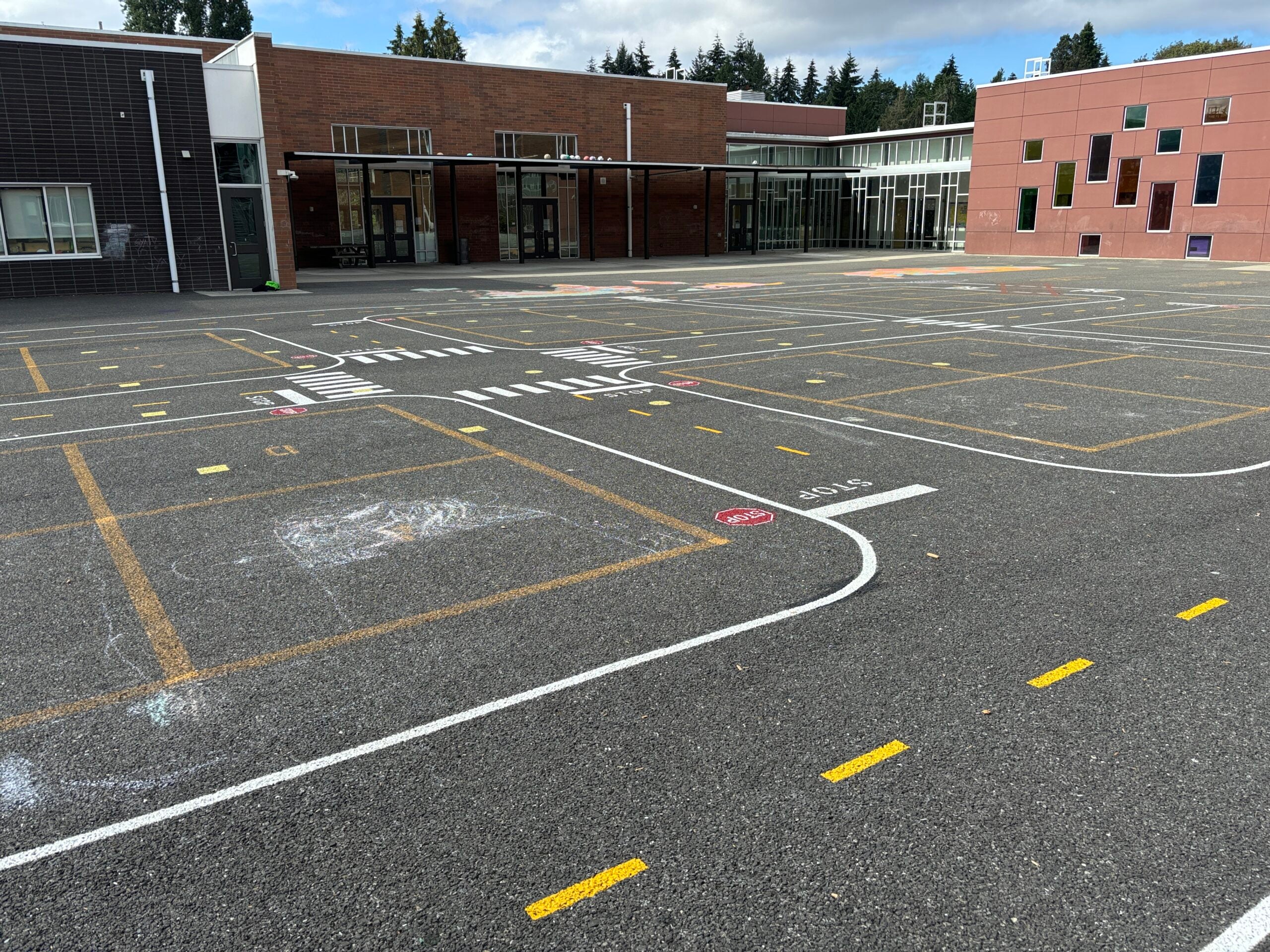 Miniature painted road curves around four square court and student chalk art. On black top with school in background.