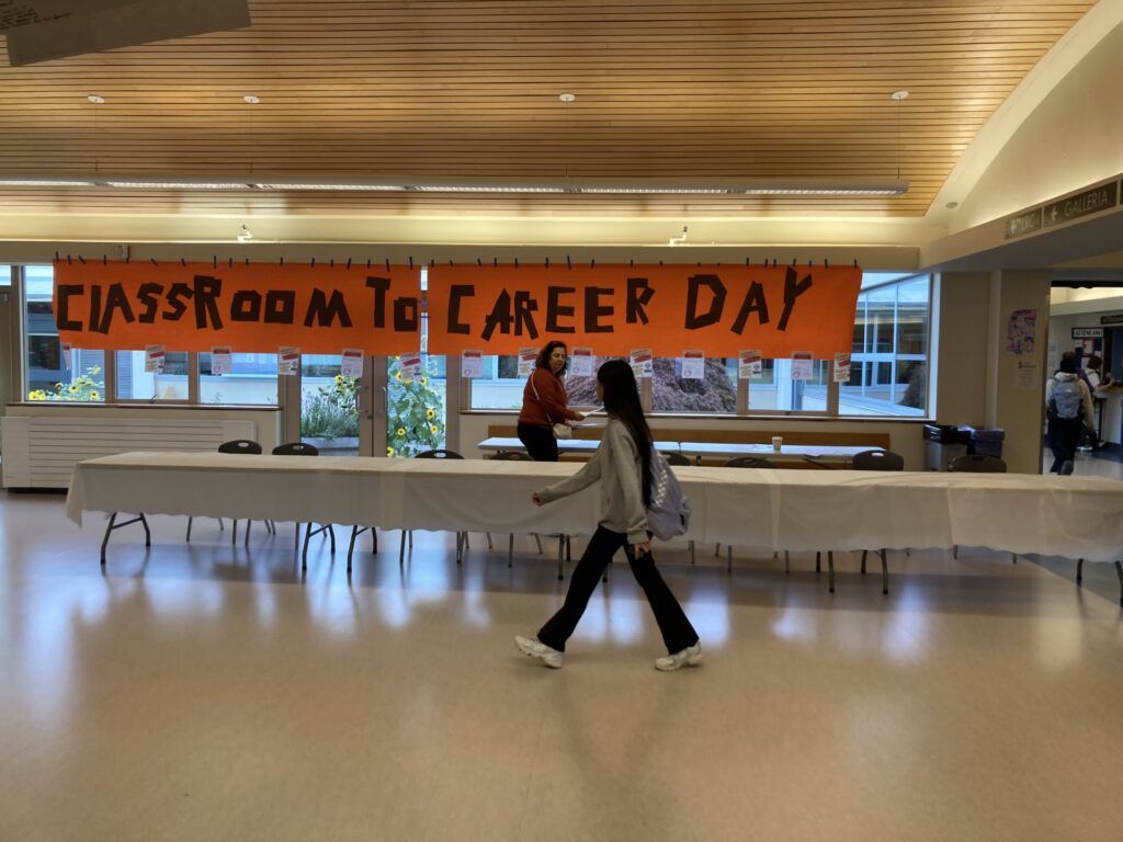 Student walking in front of set up table in front of large orange banner with words "classroom to career day" written on it. 