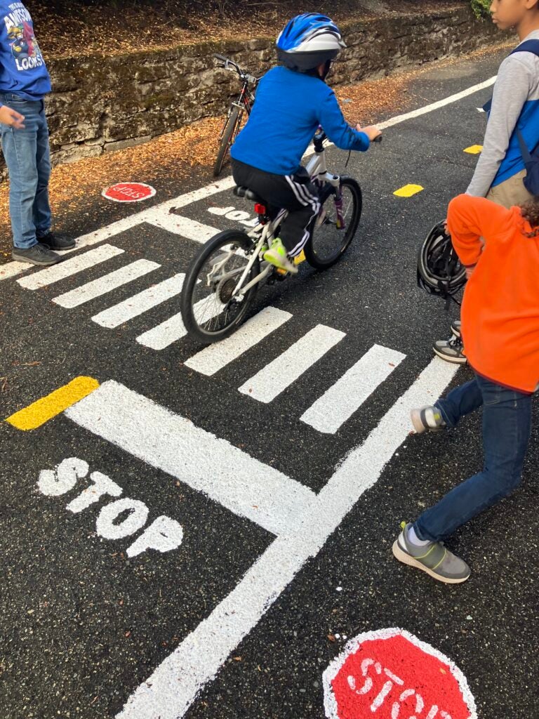 student practices biking on miniature painted street. Other students cheer them on.