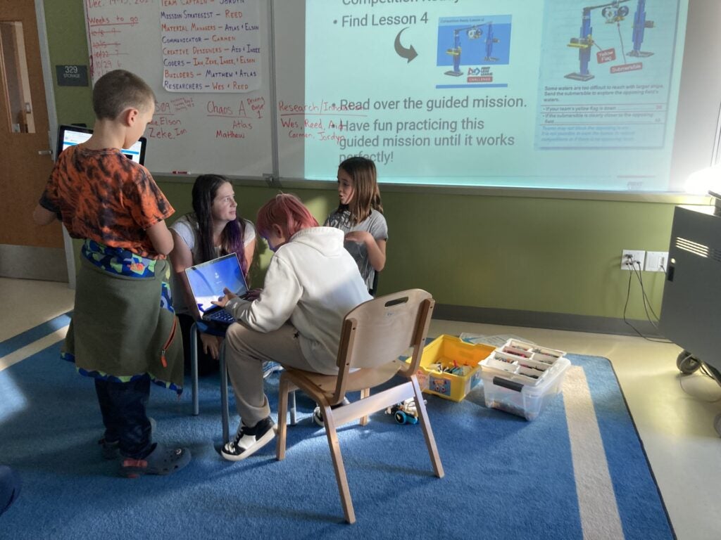 3 students gathered around teacher for robotics competition coding instruction