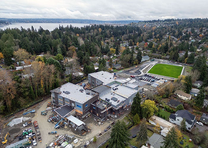 aerial of a large building under construction with a green field to the right and a lake in background