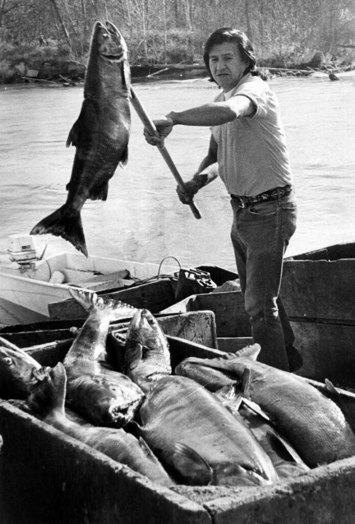 Photo of Native man holding a large salmon on a spear in a boat